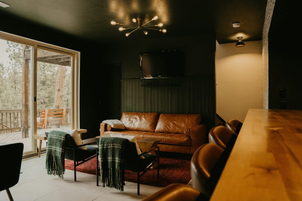 A warm, slightly blurry photo of a cosy and modern living room with a tan leather couch, dark green walls, and a wooden bar.